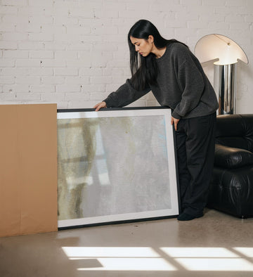 Woman holding a large mirror in a room with a white brick wall and a lamp.