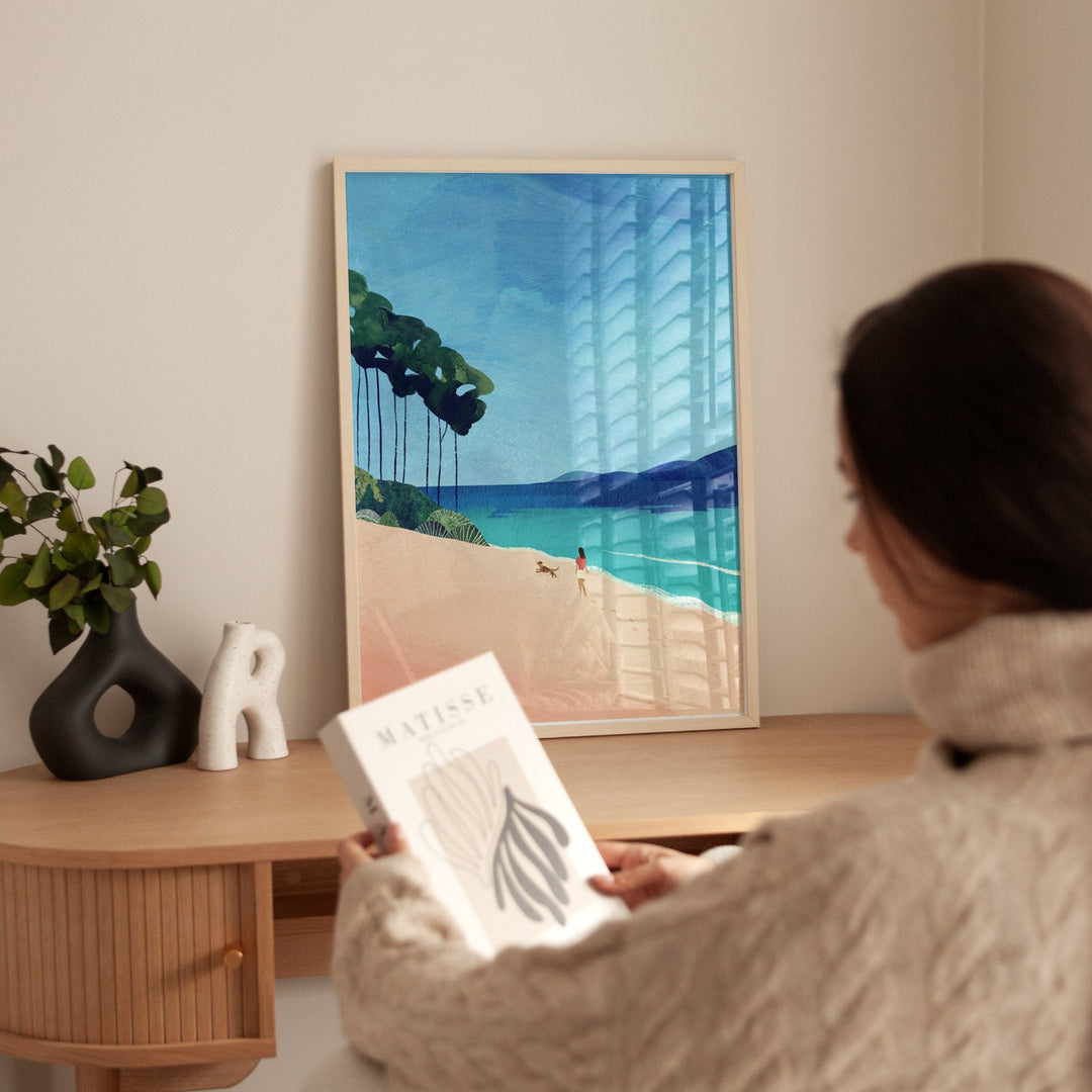 Person holding a book in front of a beach scene on a wall.