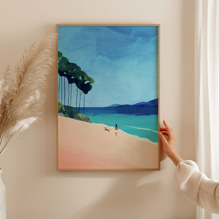 Artwork of a beach scene with a person and dog on a sand dune, held by a hand against a white wall.