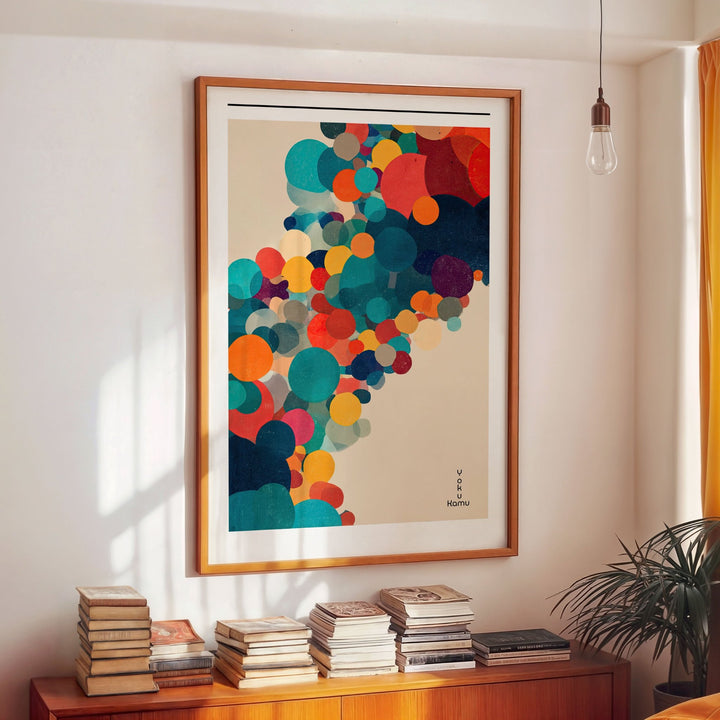 Colorful abstract art print in a wooden picture on a wall above a wooden cabinet with books.