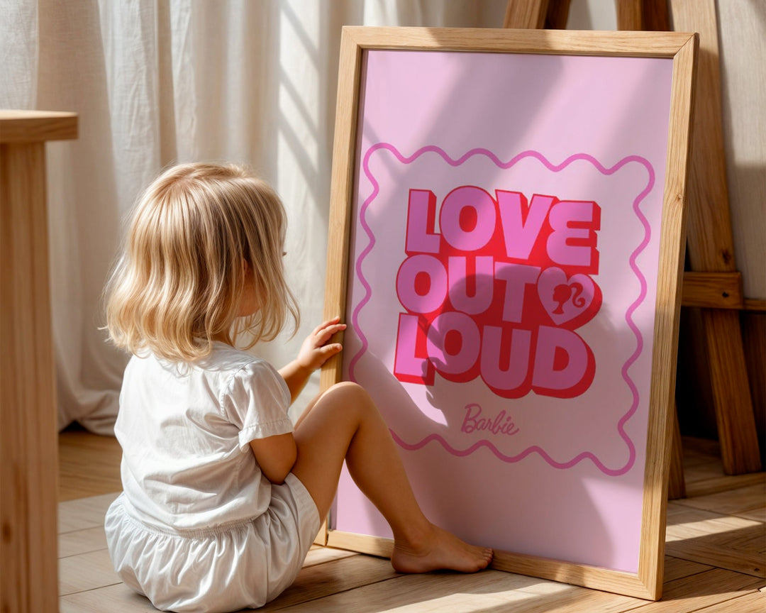 Child holding a Barbie poster with 'Love Out Loud' text on a wooden floor.