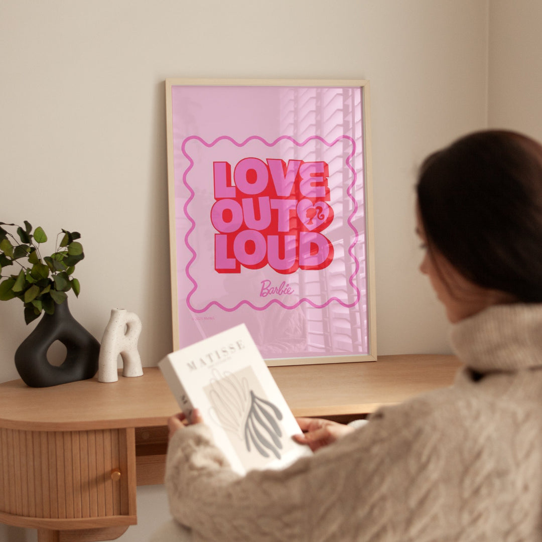 Person holding a book in front of a 'Love Out Loud' Barbie poster on a wall.
