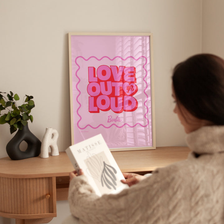 Person holding a book in front of a 'Love Out Loud' Barbie poster on a wall.