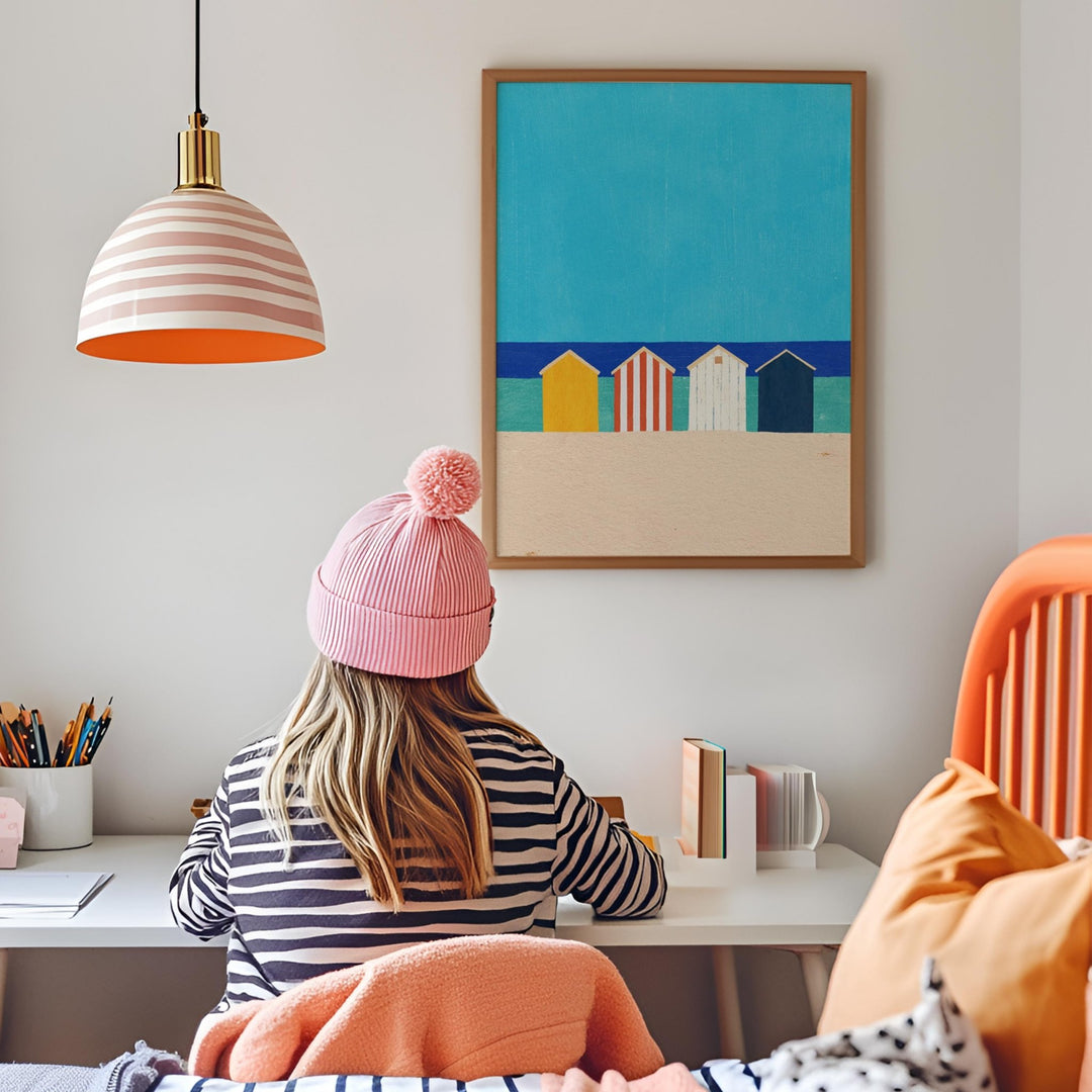 Person wearing a pink beanie sitting at a desk with a colorful painting of beach huts on the wall.