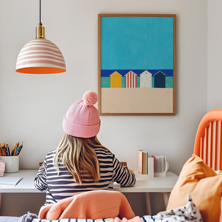 Person wearing a pink beanie sitting at a desk with a colorful painting of beach huts on the wall.