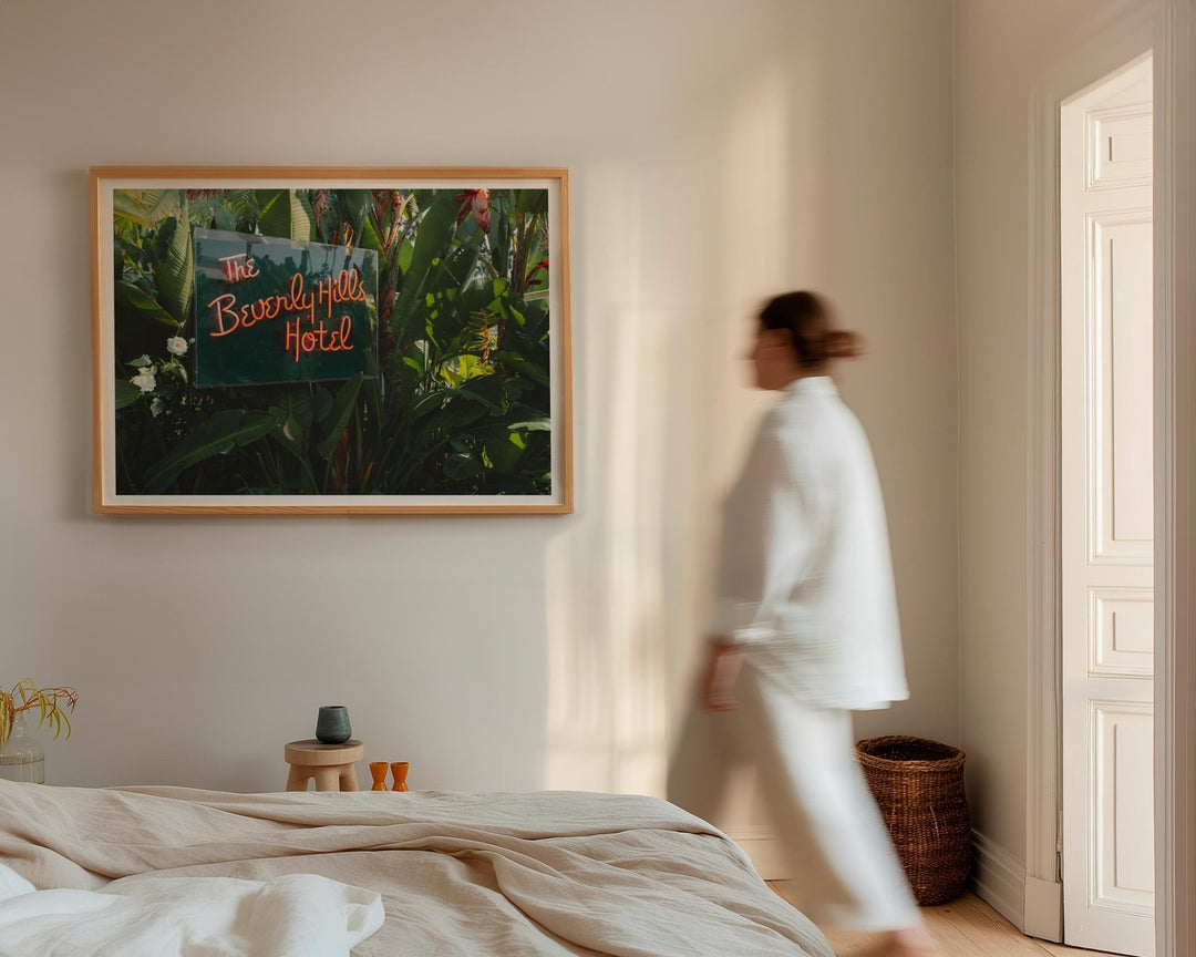 Person in a white robe walking in a bedroom with a picture of the Beverly Hills Hotel on the wall.