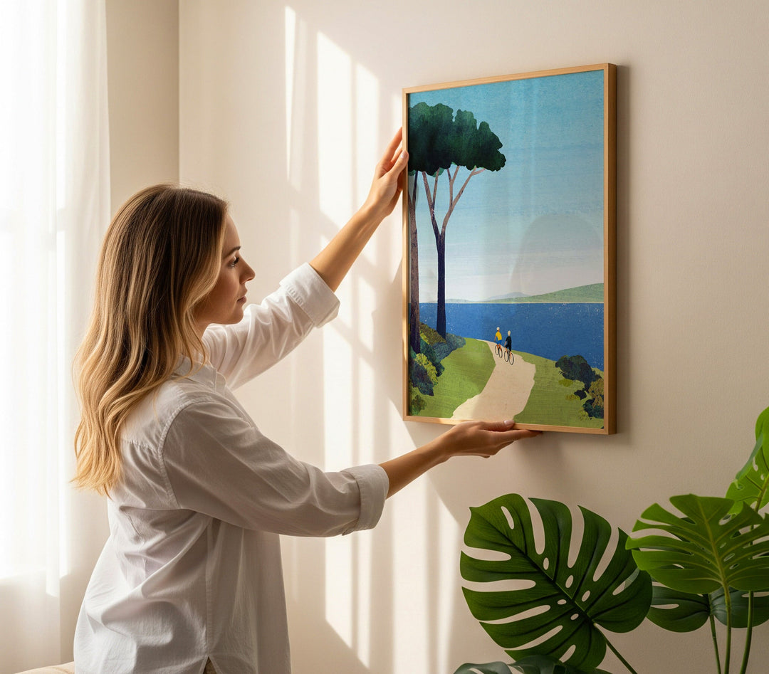 Woman hanging an artwork of a landscape with trees and a path on a wall.