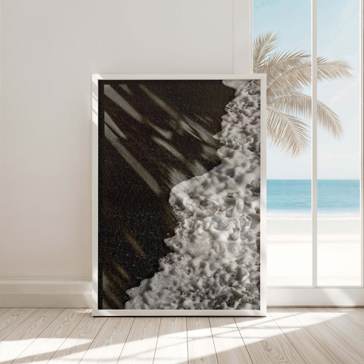 Artwork of a beach scene with waves and palm tree, placed in a room with a window view of the ocean.