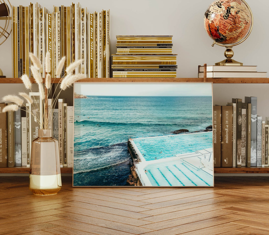 artwork of a pool and ocean scene on a wooden shelf with books and decor.