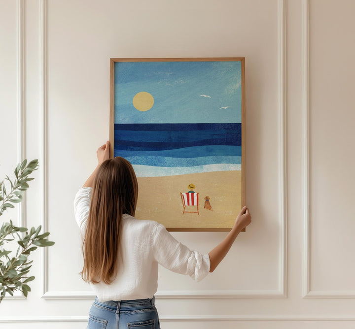 Person hanging an artwork of a beach scene on a wall.
