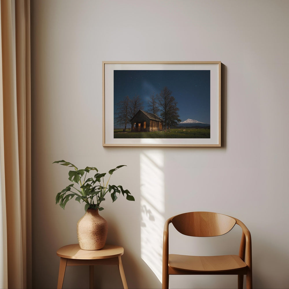 Artwork of a cabin scene on a wall with a plant and chair below.