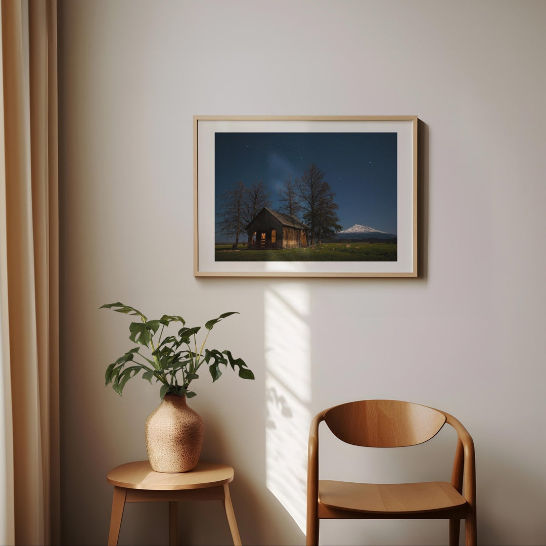 Artwork of a cabin scene on a wall with a plant and chair below.