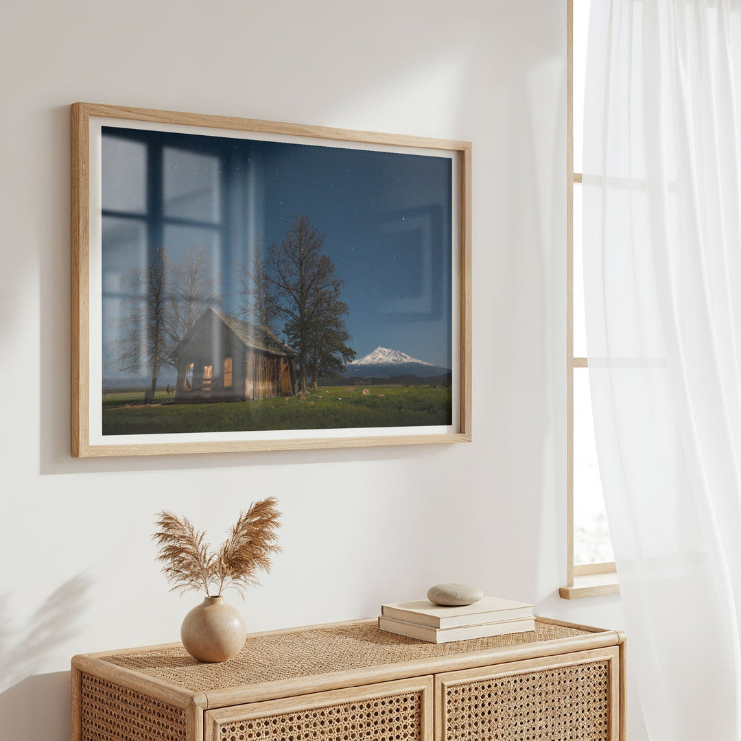 Artwork of a landscape with a house and mountain on a wall in a room with a wicker console table.