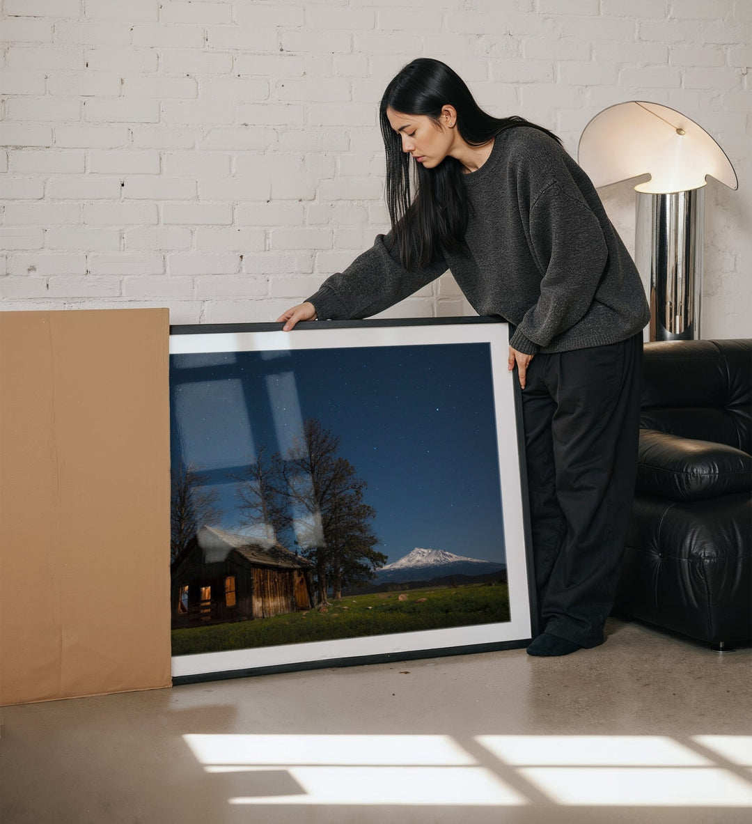 Woman holding a large picture of a landscape with a mountain in a living room.