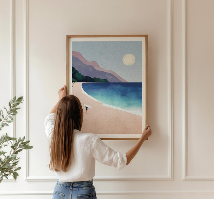 Woman hanging artwork of a beach scene on a wall.