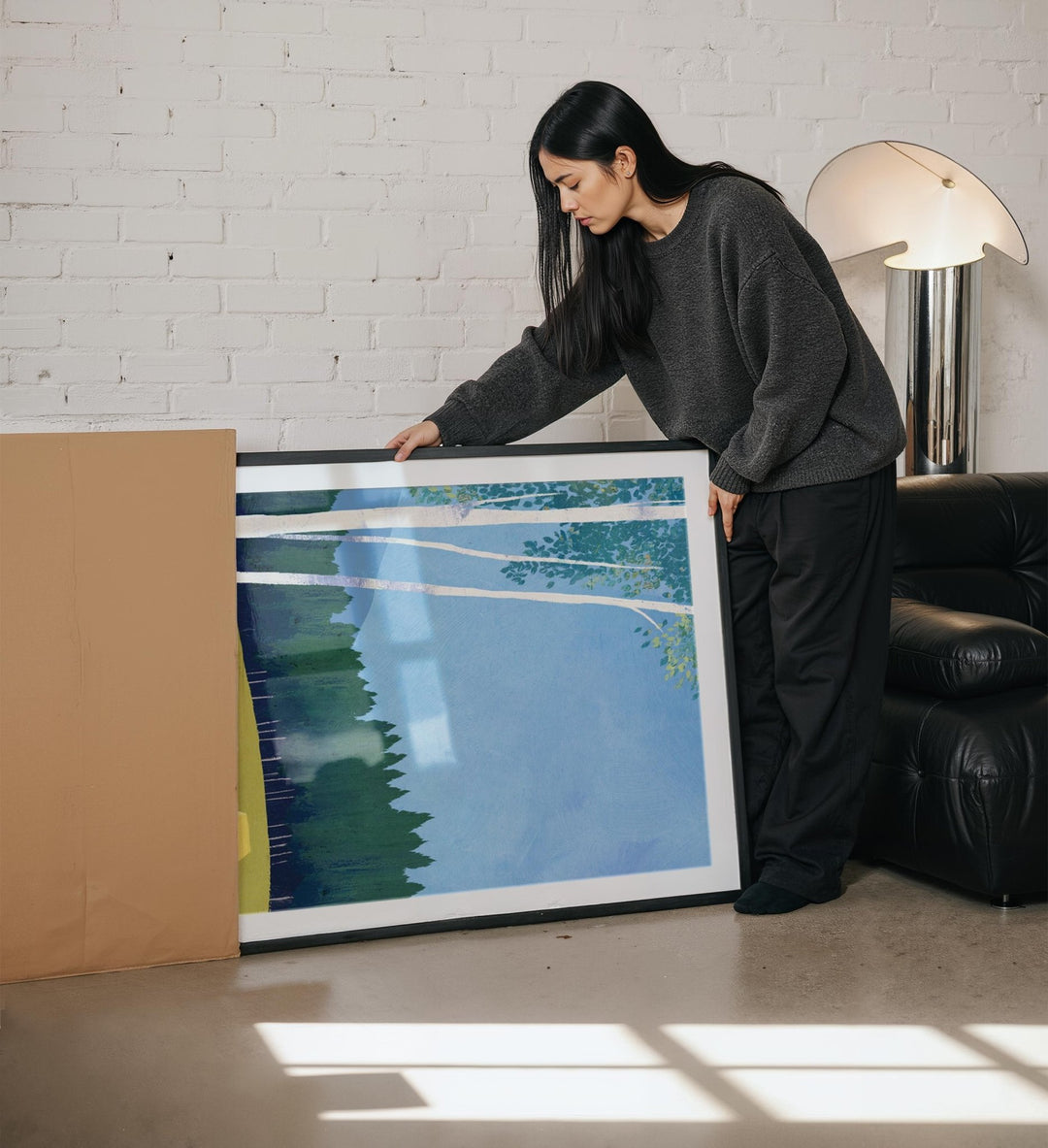 Woman holding a large artwork in a room with a white brick wall and a lamp.
