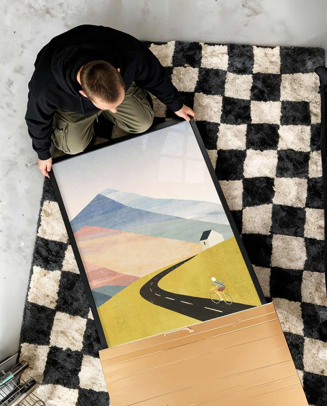 Person unboxing a artwork of a cyclist on a road with mountains, on a checkered rug.