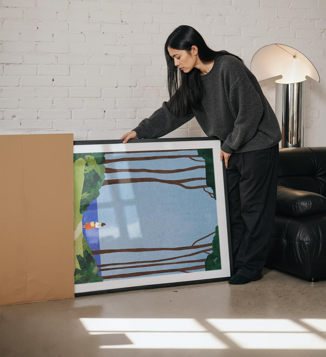 Woman holding a painting in a room with a white brick wall and a lamp.