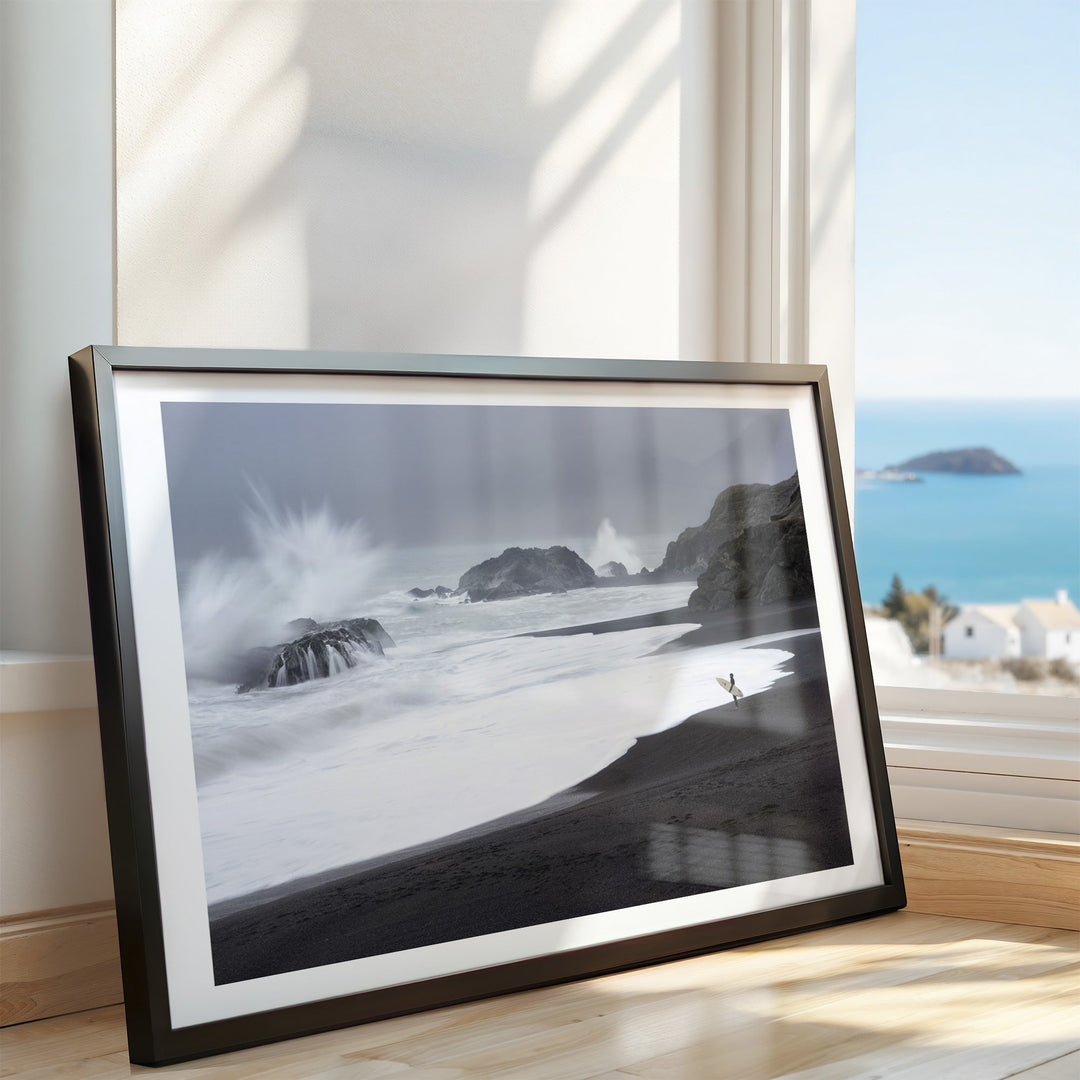 Artwork of a coastal scene with waves and rocks, placed on a surface with a window view in the background.