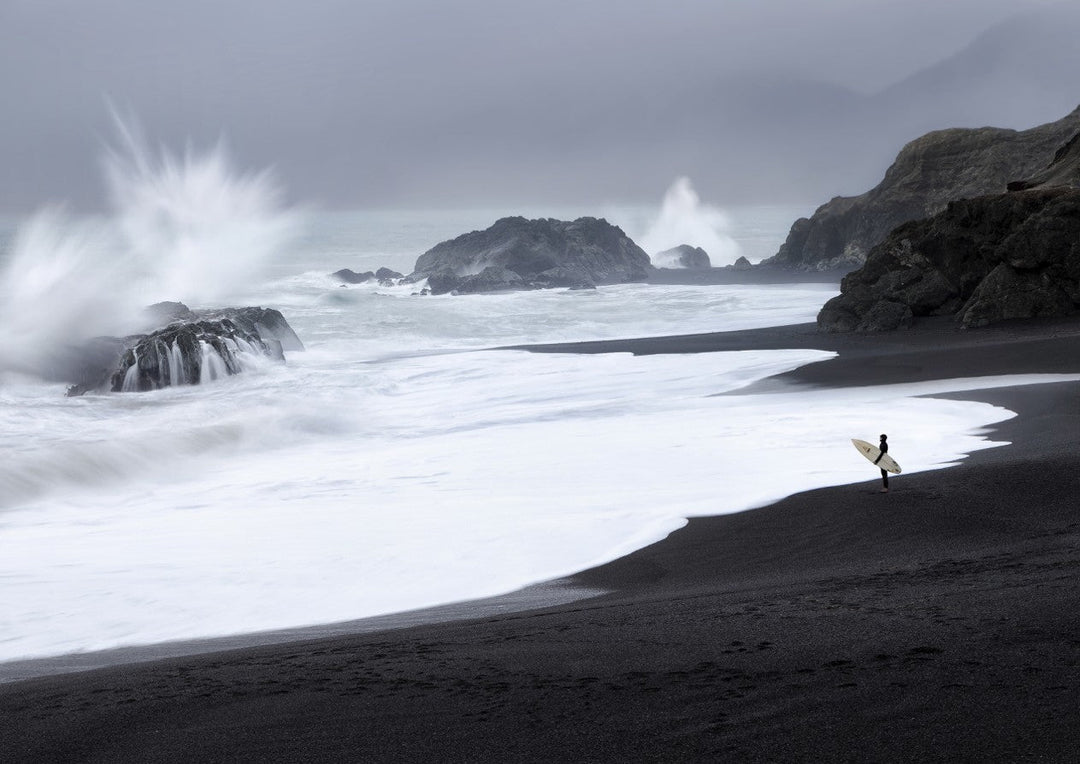 A lone surfer stands on a black sand beach, holding a surfboard and gazing at large waves crashing against rocky outcrops under a cloudy, gray sky. framed