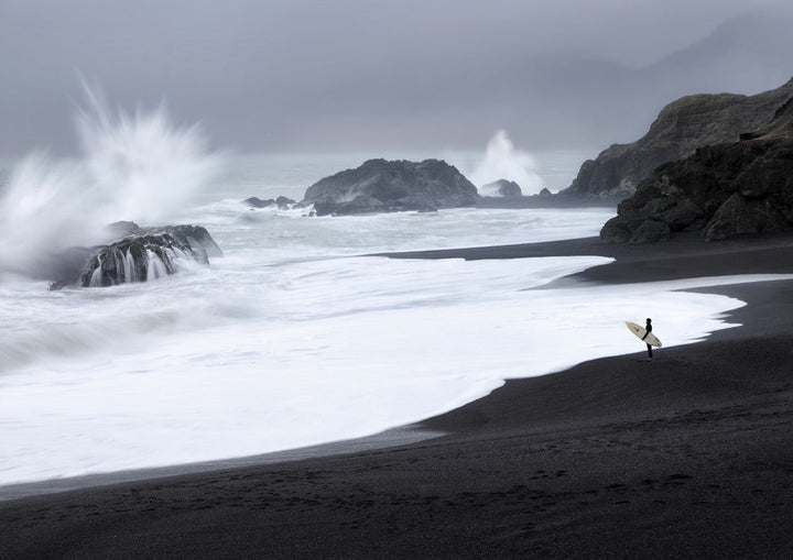 A lone surfer stands on a black sand beach, holding a surfboard and gazing at large waves crashing against rocky outcrops under a cloudy, gray sky. framed