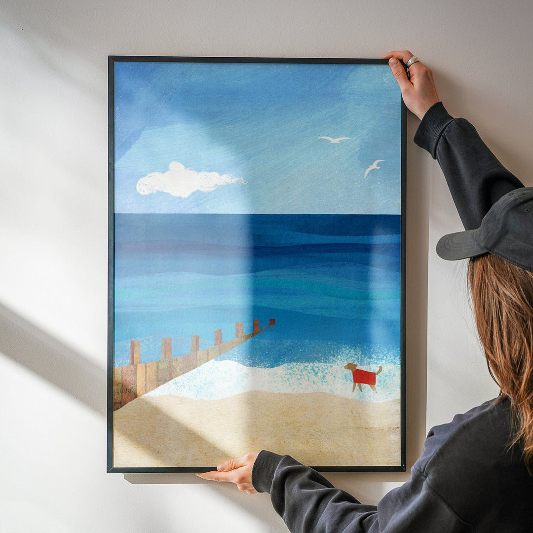 Person hanging an artwork of a beach scene with a dog on a white wall.