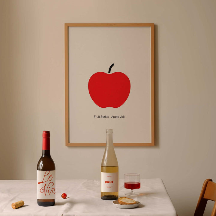 Dining table with wine bottles, a glass of red wine, and a picture of an apple on a beige wall.