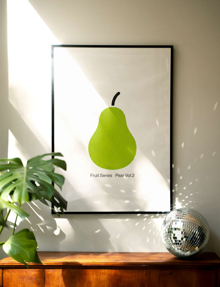 artwork of a green pear on a wall with a plant and decorative ball in the foreground.