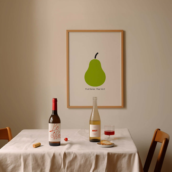 Dining table with wine bottles, a glass of red wine, and a picture of a green pear on a beige wall.