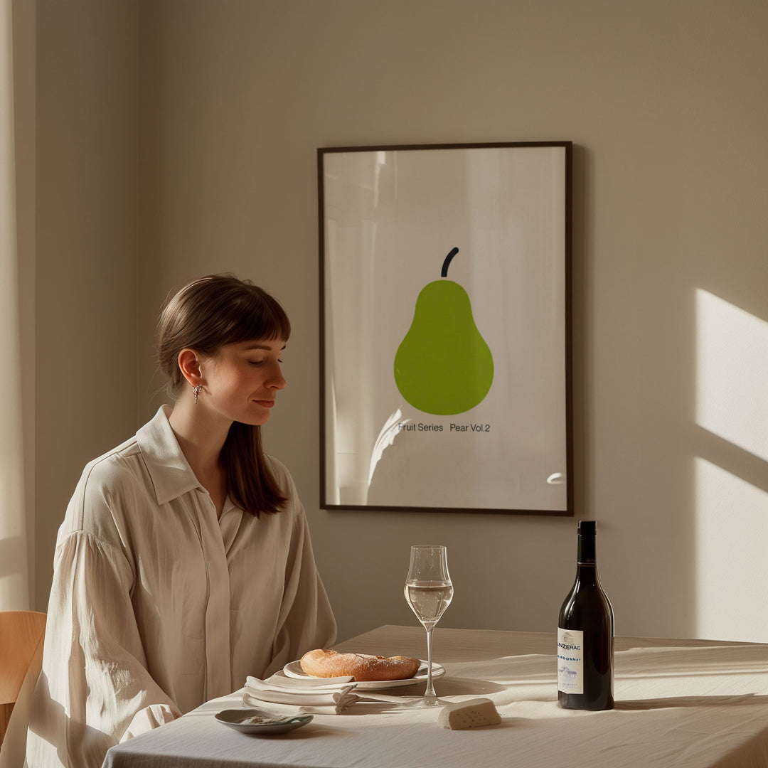 Woman sitting at a table with a glass of wine and a bottle, in front of a wall with a green pear artwork.