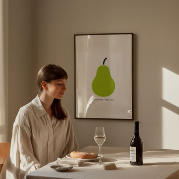 Woman sitting at a table with a glass of wine and a bottle, in front of a wall with a green pear artwork.