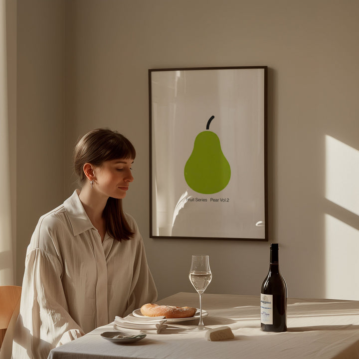 Woman sitting at a table with a glass of wine and a bottle, in front of a wall with a green pear artwork.