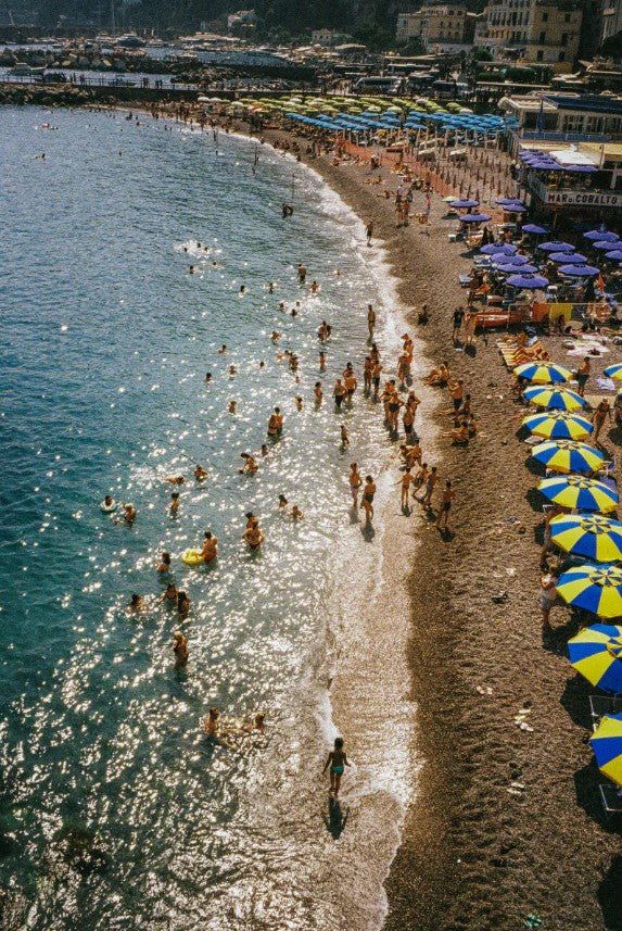 An overhead, sun-drenched photograph captures a crowded, crescent-shaped Mediterranean pebble beach with many people swimming in the sparkling blue water and lounging beneath rows of blue and yellow striped umbrellas, with a seaside town visible in the background. framed