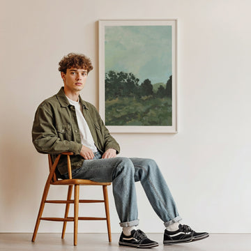 Man sitting on a wooden stool in a room with a landscape painting on the wall.