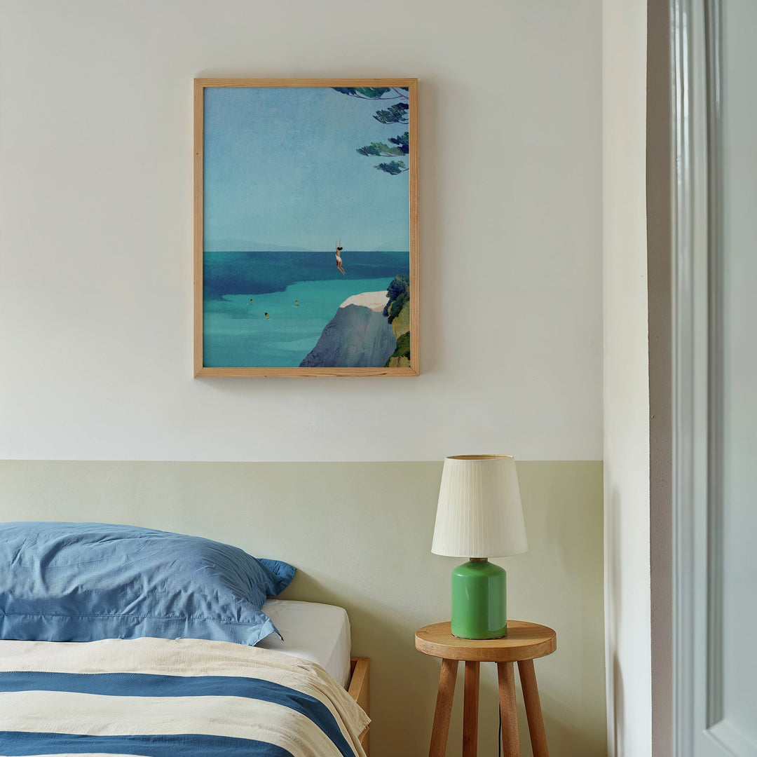 Bedroom with a painting of a beach scene on the wall, a green lamp on a wooden side table, and blue bedding.