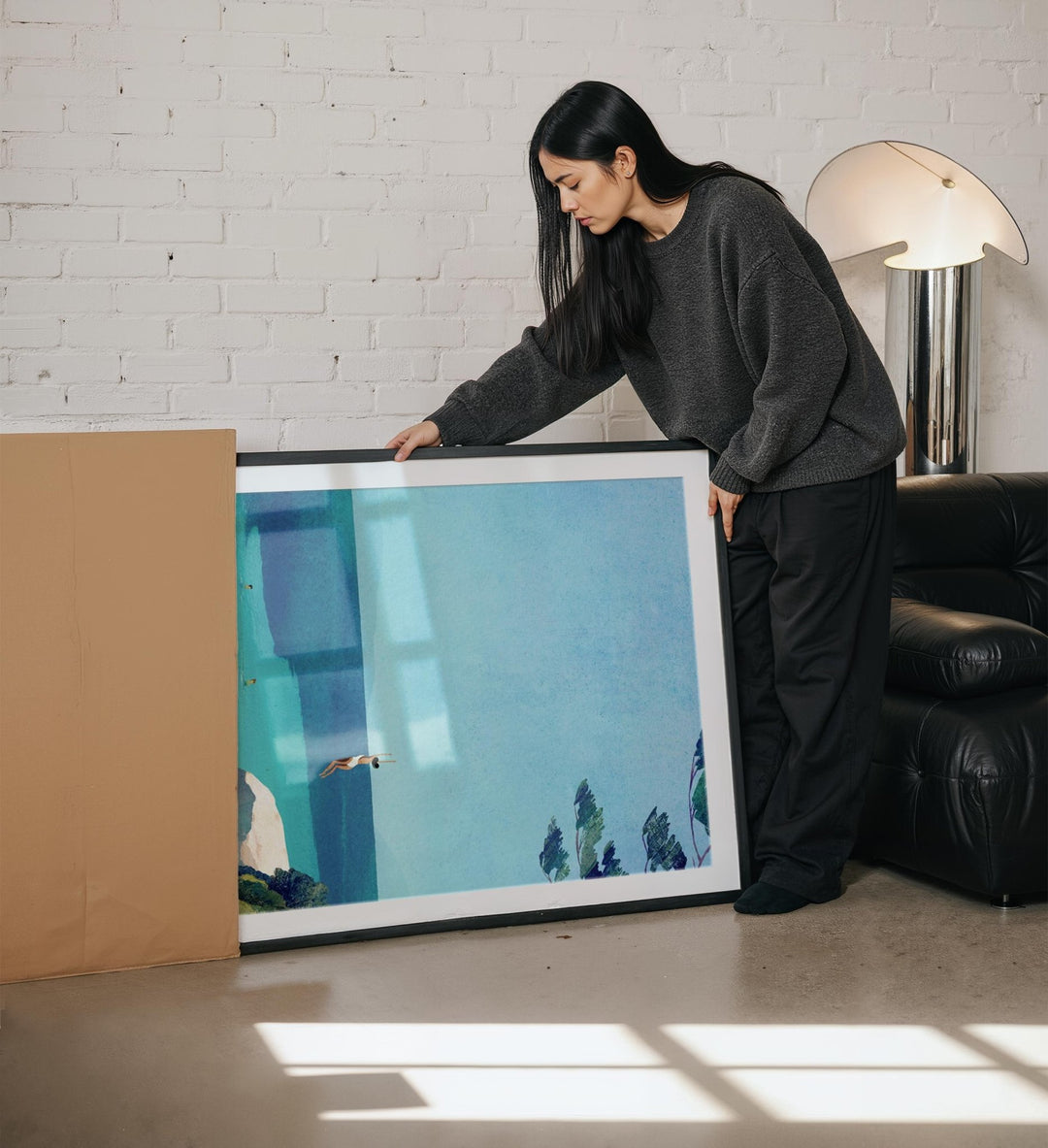Woman holding a large artwork in a room with a white brick wall and a lamp.