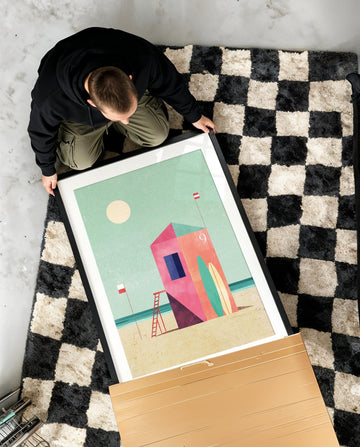 Person holding an artwork of a colorful beach scene on a checkered rug.