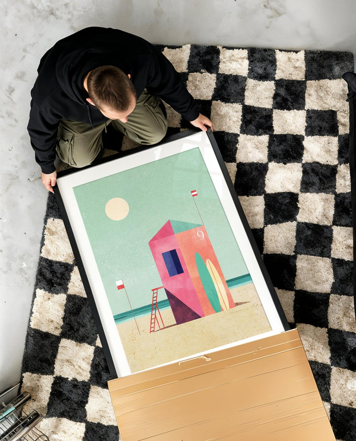 Person holding an artwork of a colorful beach scene on a checkered rug.