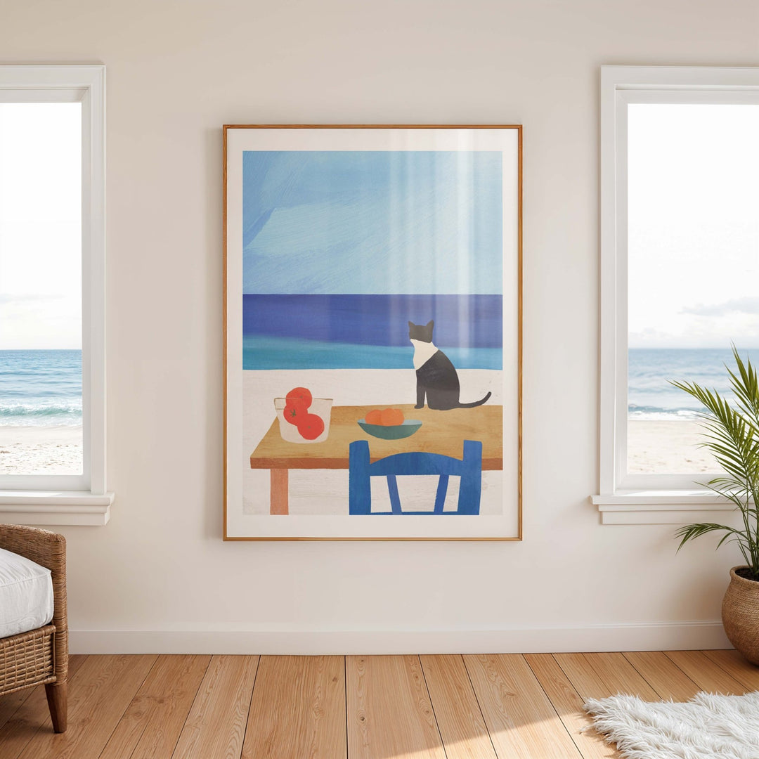 Artwork of a cat at a table with a beach scene, hanging on a wall with large windows.