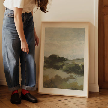 Person wearing blue jeans and red socks standing next to a landscape painting on a wooden floor.