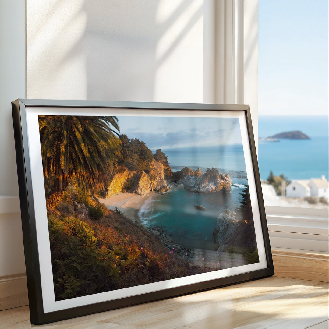 Artwork of a coastal scene with a body of water and trees, placed on a surface with a window view in the background.