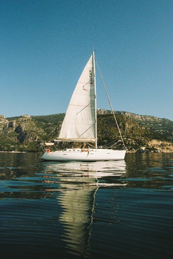 A photograph shows a white sailboat with a tall mainsail floating on dark, calm water, with its reflection visible against a backdrop of a steep, green, rocky hillside under a clear blue sky. framed