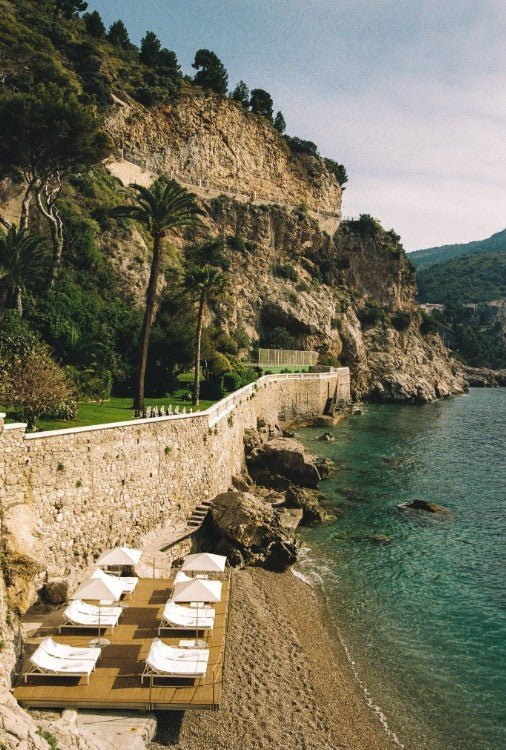 A sunlit photograph captures a luxurious Mediterranean hideaway featuring white sun loungers on a wooden deck next to a small pebble beach and clear turquoise water, all overlooked by towering, rugged stone cliffs and a high retaining wall. framed