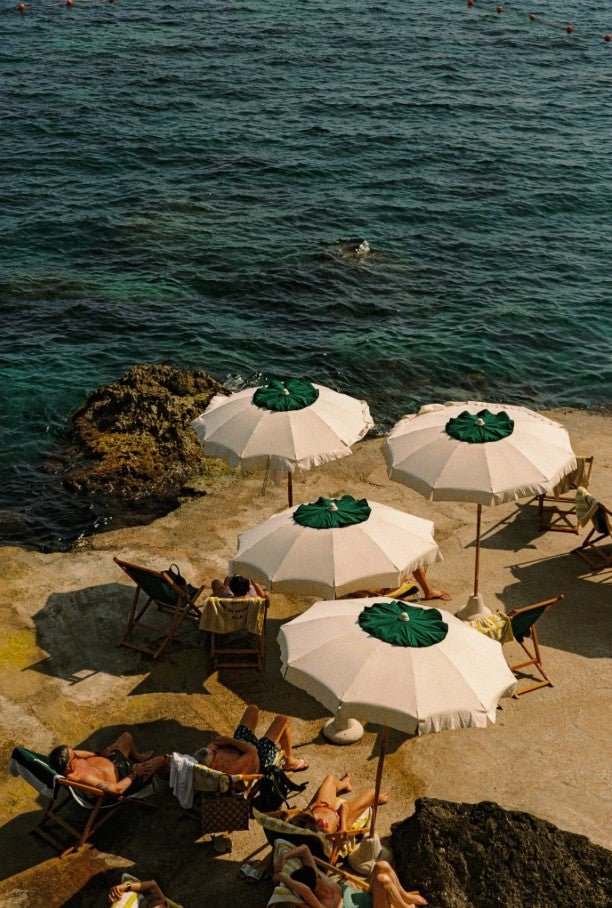 An elevated, sunlit photograph captures a Mediterranean swimming spot where people are sunbathing on deck chairs on a concrete ledge next to dark teal water, shaded by four large, white parasols with contrasting green centers. frame