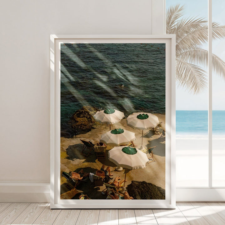 print, leaning against a white wall and floor with wooden planks, displays an overhead photo of sunbathers relaxing under four white and green parasols on a concrete ledge next to dark blue Mediterranean water.