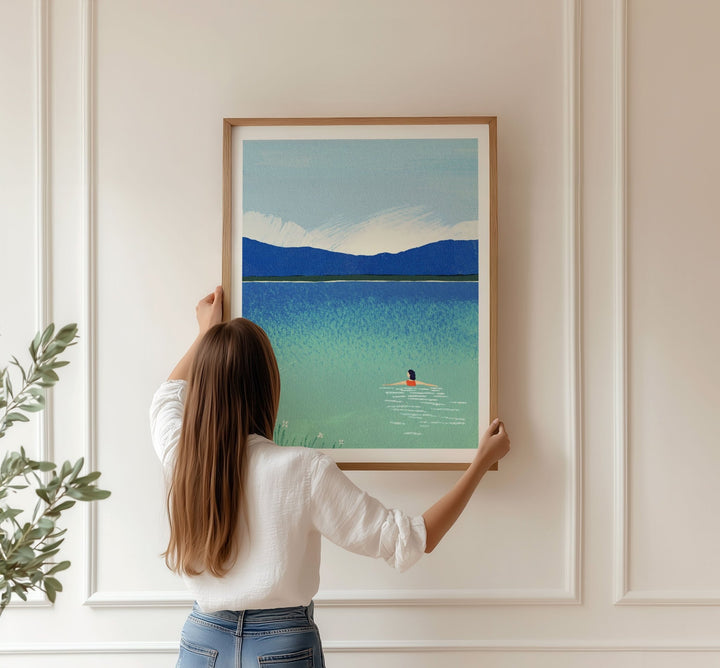Person hanging an artwork of a lake scene on a wall.