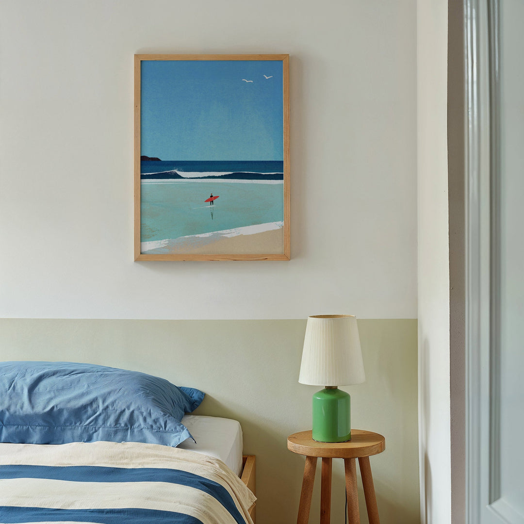 Artwork of a beach scene above a bed with a green lamp on a side table.