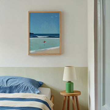 Artwork of a beach scene above a bed with a green lamp on a side table.