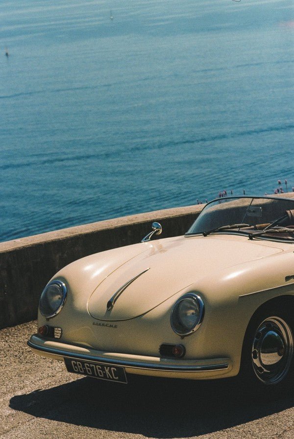A vintage, pale yellow Porsche convertible is photographed in a close-up, high-angle shot, parked along a concrete wall overlooking the vast, deep blue ocean. framed