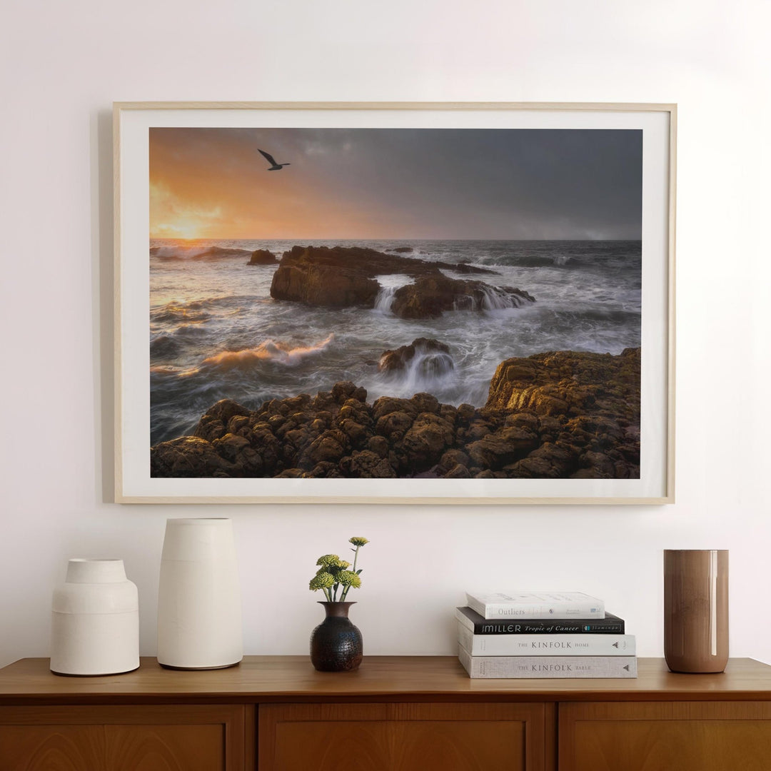 Artwork of a sunset over rocky ocean scene above a wooden cabinet with decorative items.